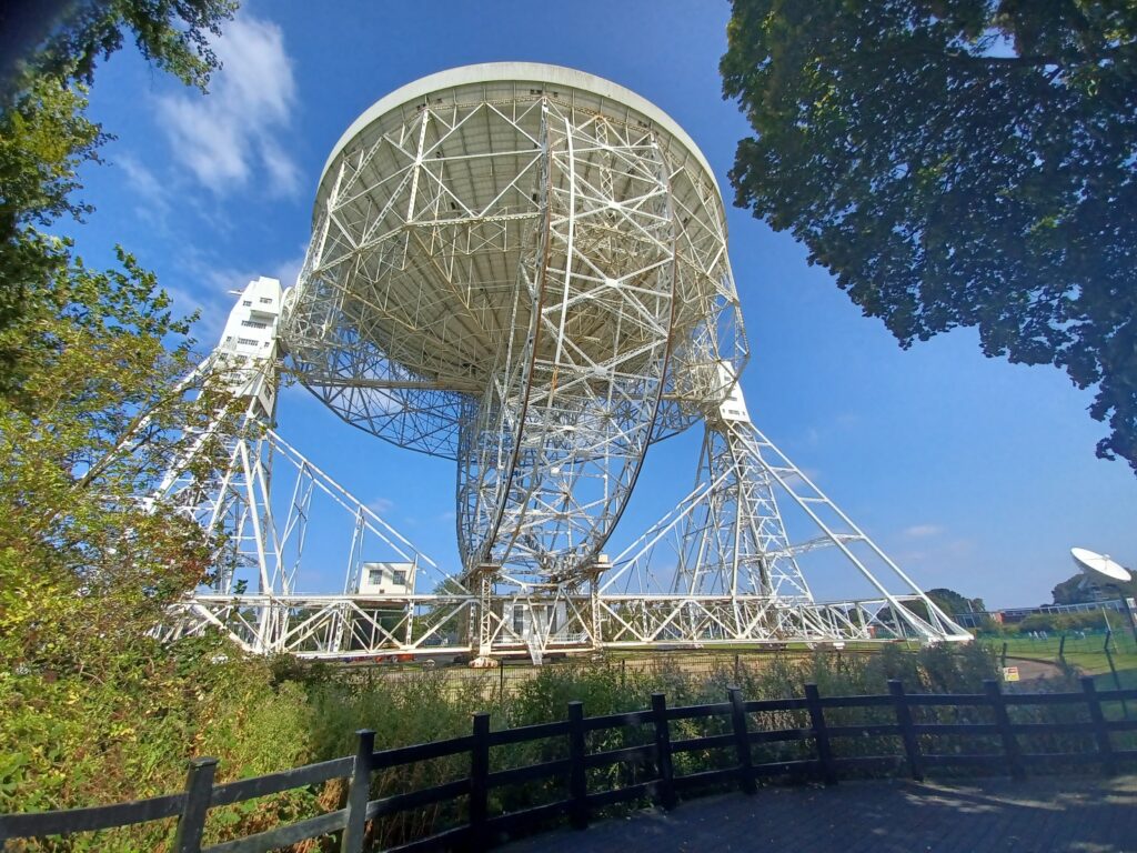 The Mk I radio telescope at Jodrell Bank, pictured through trees against a clear blue sky.