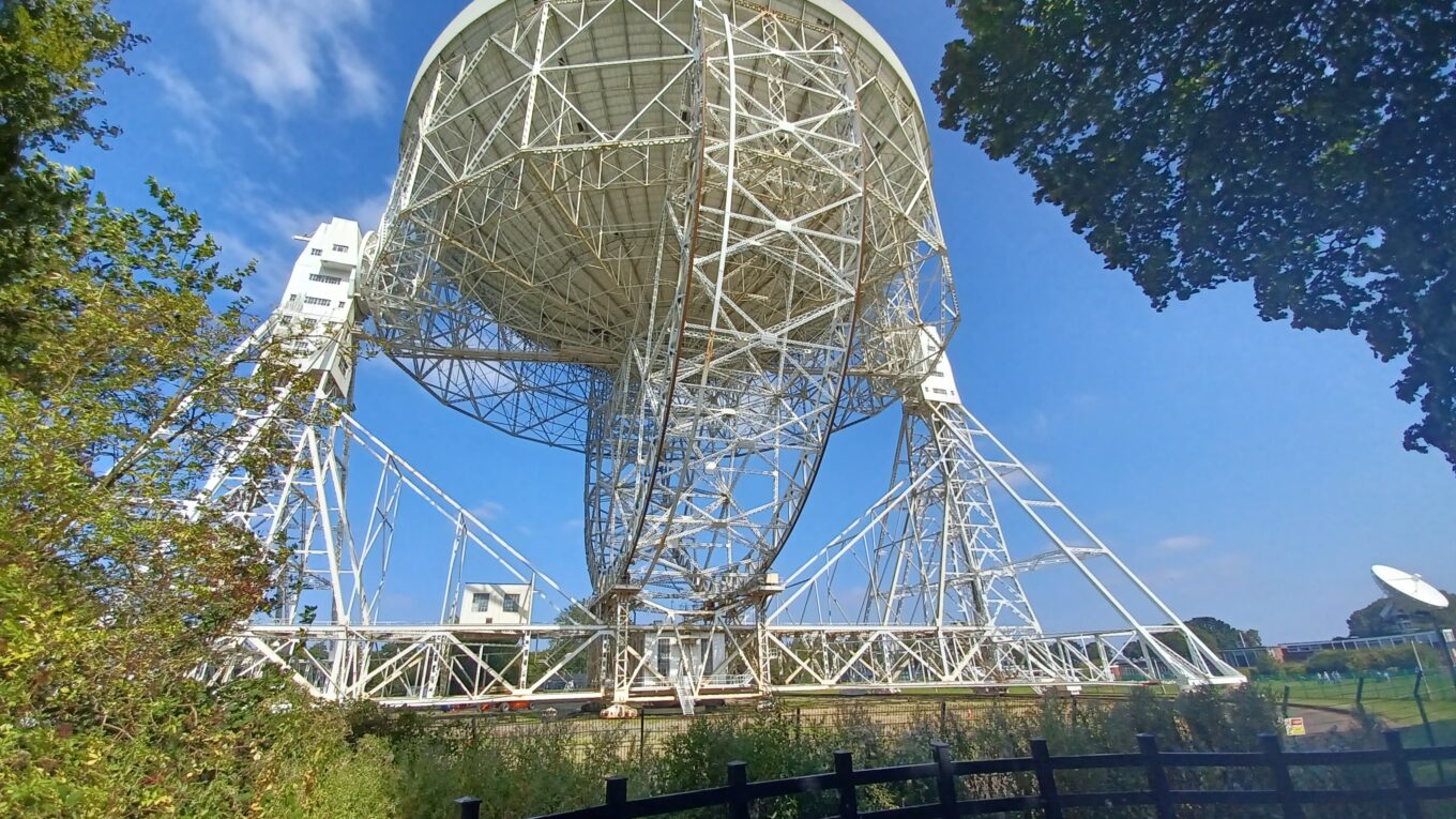 The Mk I radio telescope at Jodrell Bank, pictured through trees against a clear blue sky.